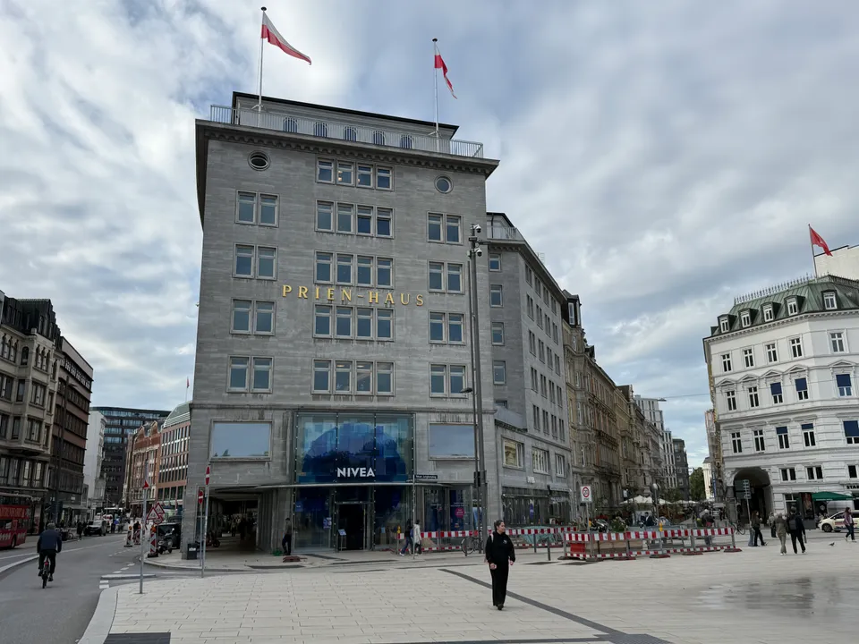 Hellgraues Eckgebäude mit goldener Aufschrift Prien-Haus, Fahnen auf dem Dach und verglastem NIVEA-Eingang am Platz