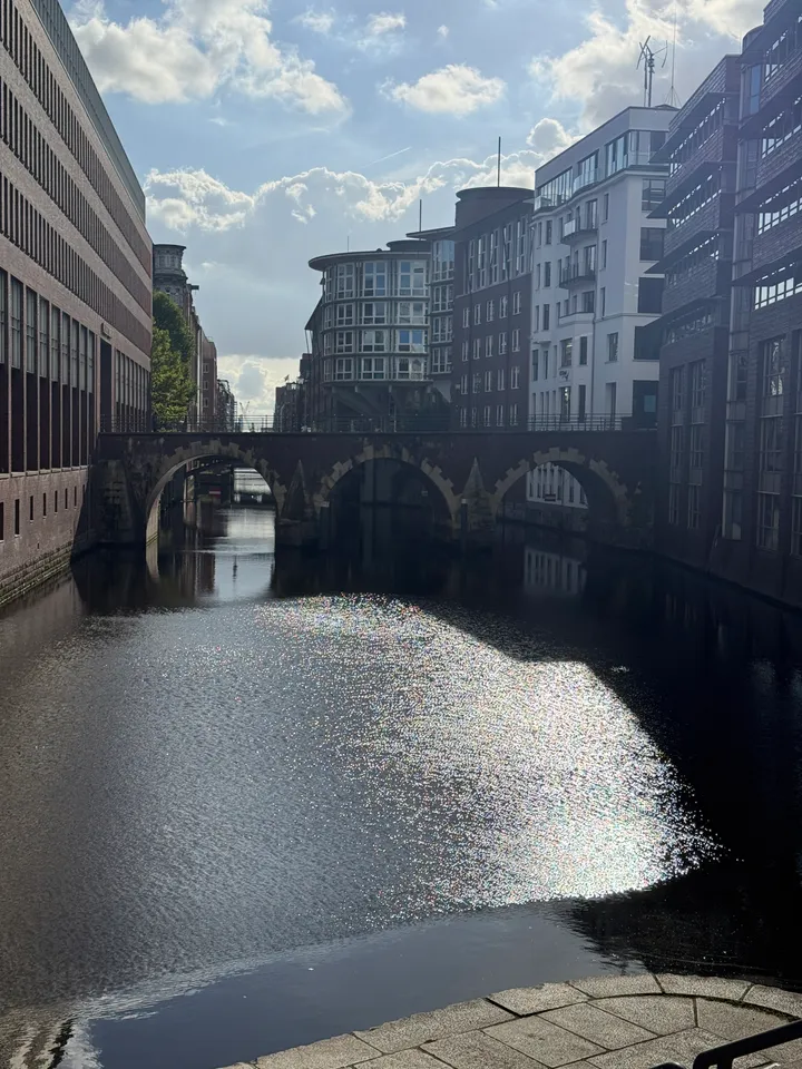 Fleet zwischen Backstein- und Glasgebäuden mit dreibogiger Brücke im Gegenlicht, Sonnenlicht glitzert auf dem Wasser