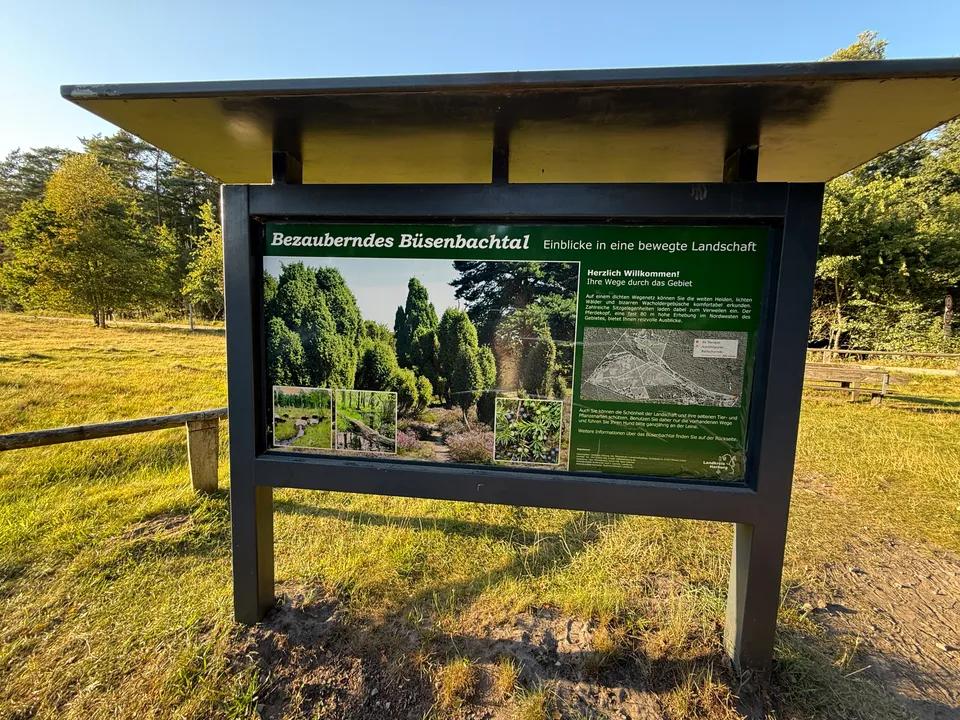 Überdachte Infotafel „Bezauberndes Büsenbachtal“ mit Landschaftsfotos und Karte auf sonniger Wiese vor Bäumen