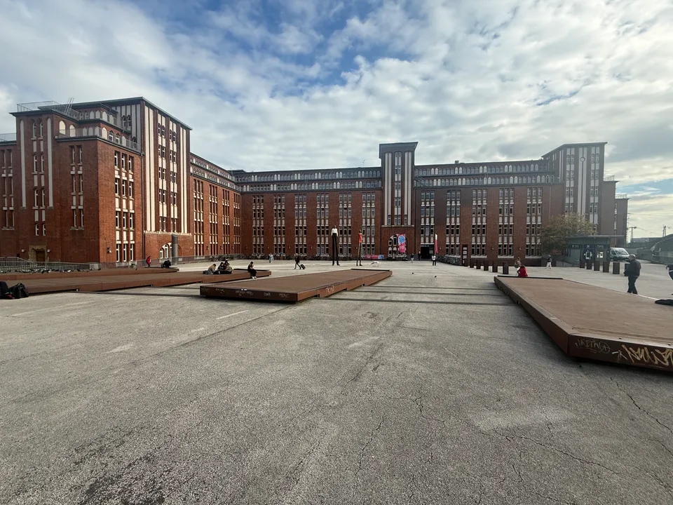 Großer Platz mit rostbraunen Sitzpodesten vor mehrflügeligem Backsteingebäude mit Türmen unter bewölktem Himmel