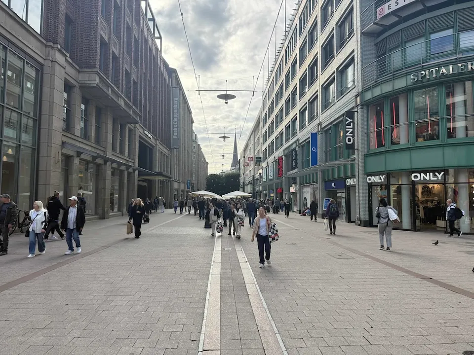 Belebte Fußgängerzone mit Geschäftshäusern, Passanten, Hängeleuchten über der Straße und Kirchturm im Hintergrund