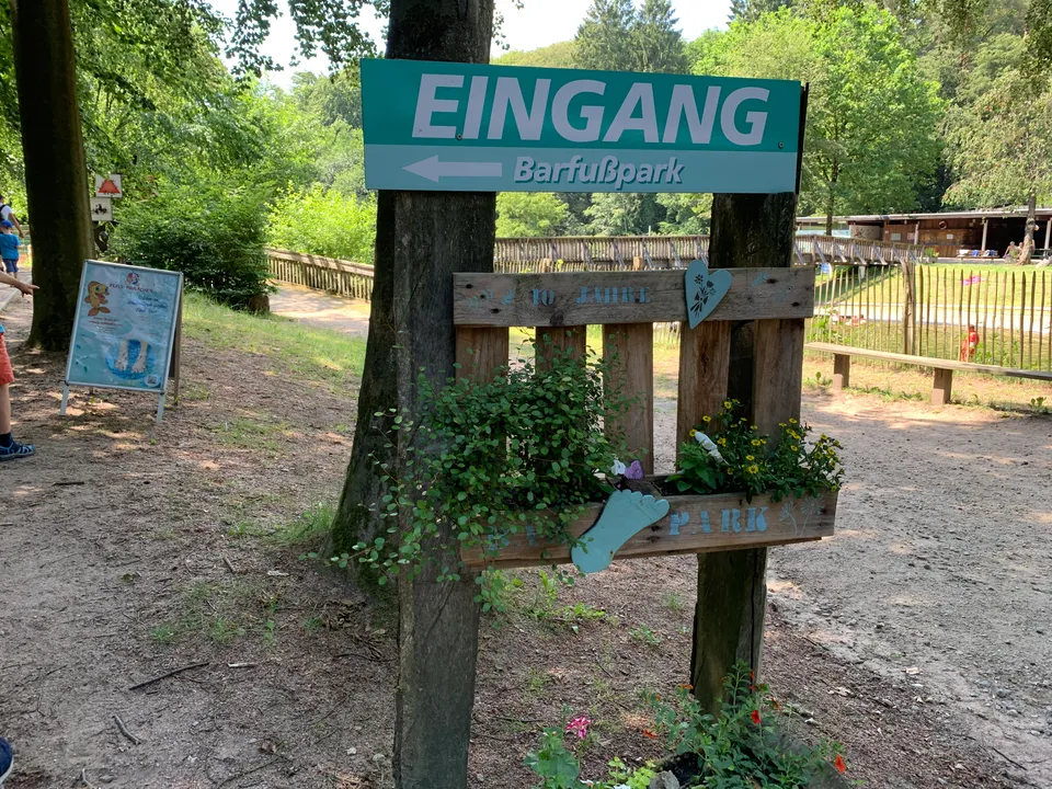 Türkisfarbener Wegweiser „Eingang Barfußpark“ an Holzpfosten, darunter bepflanzte Holzpalette mit Fußabdruck-Dekoration