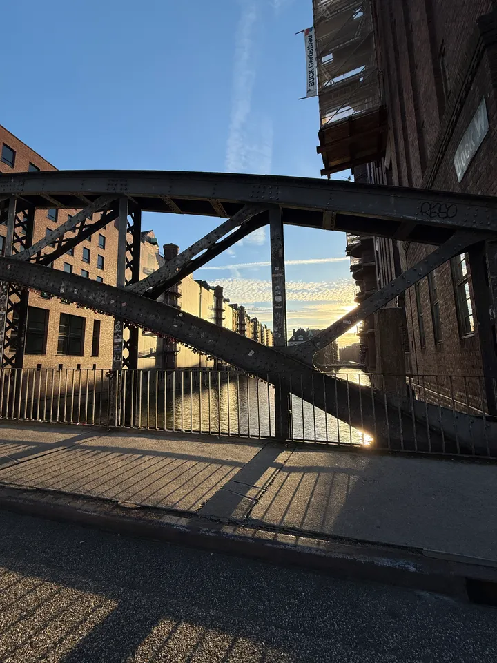 Genietete Stahlstreben einer Bogenbrücke, dahinter Fleet, Speichergebäude und tiefstehende Sonne, lange Schatten auf dem Gehweg