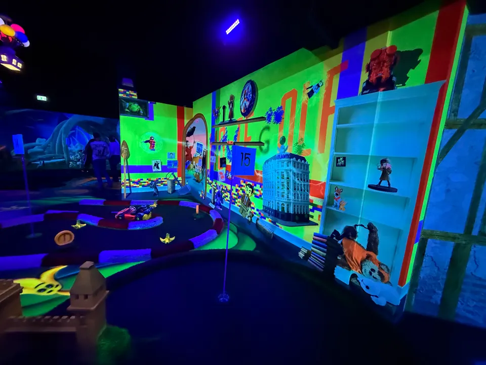 Minigolfbahn 15 vor bunter Kinderzimmerkulisse mit Regal, daneben eine Bahn im Rennstrecken-Design mit Spielzeugautos