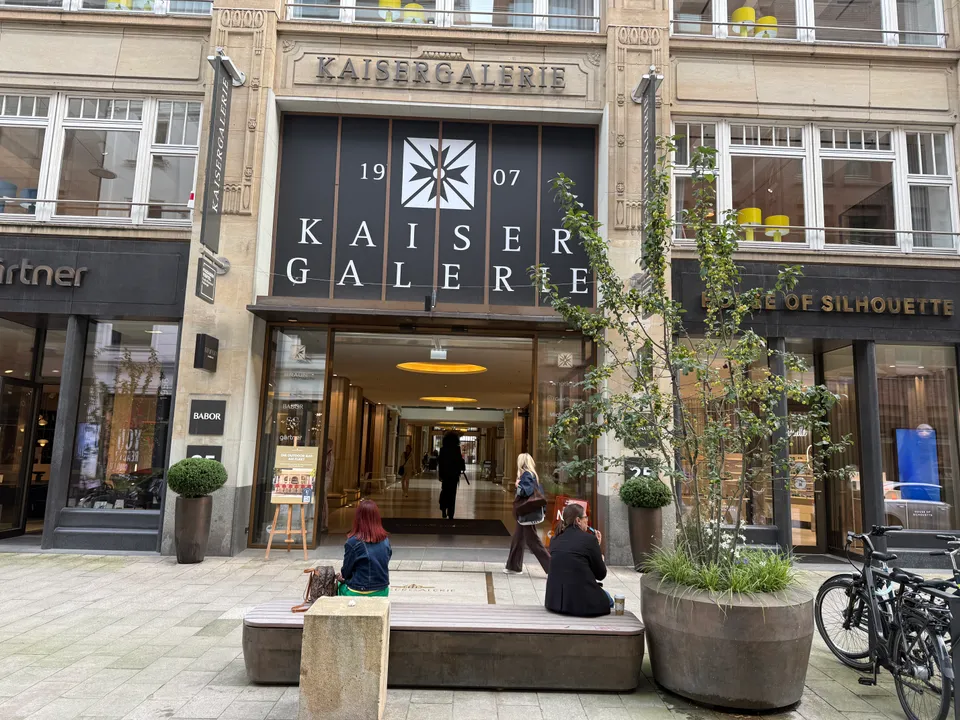Eingang der Kaisergalerie mit dunklem Banner über Glastür, davor Sitzbank, Pflanzkübel und Fahrräder
