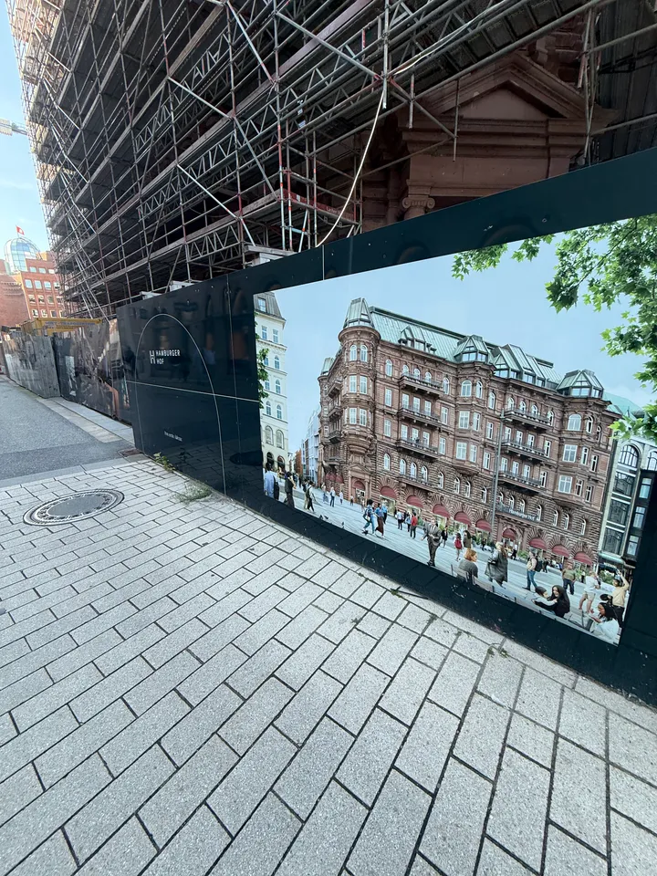 Bauzaun mit Gebäudevisualisierung vor eingerüstetem Sandsteinbau, davor ein breiter gepflasterter Gehweg