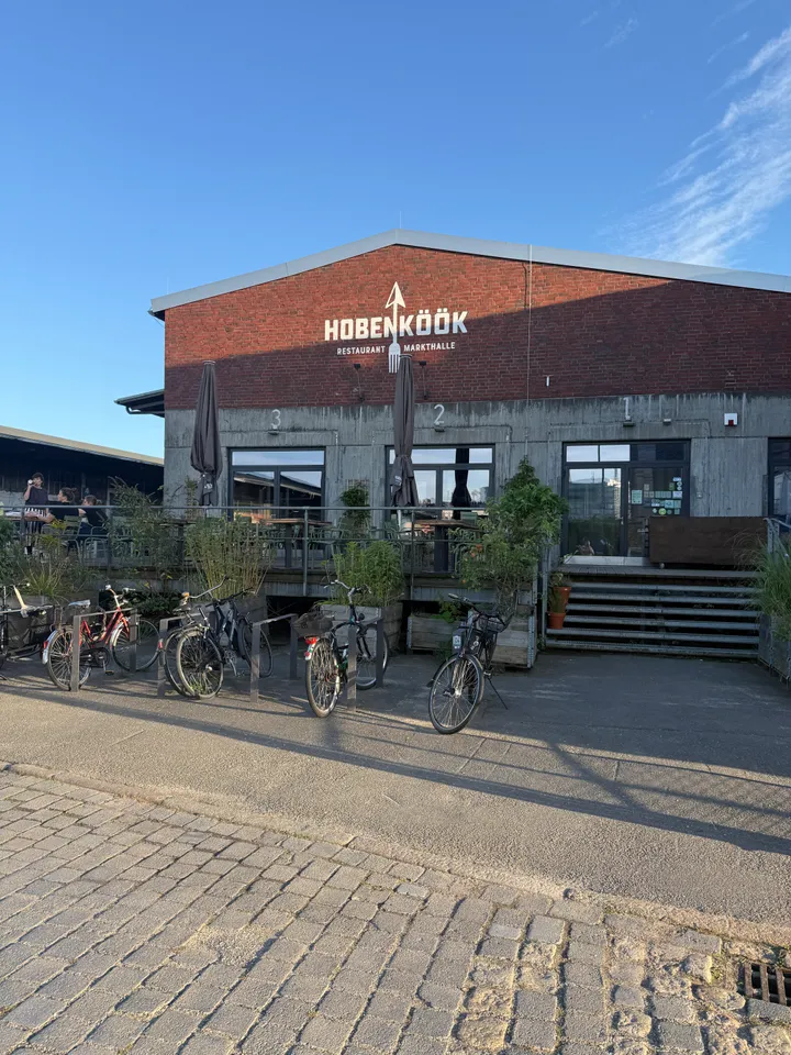 Backsteinhalle mit Schriftzug Hobenköök Restaurant Markthalle, davor Terrasse mit Pflanzen und abgestellten Fahrrädern bei blauem Himmel